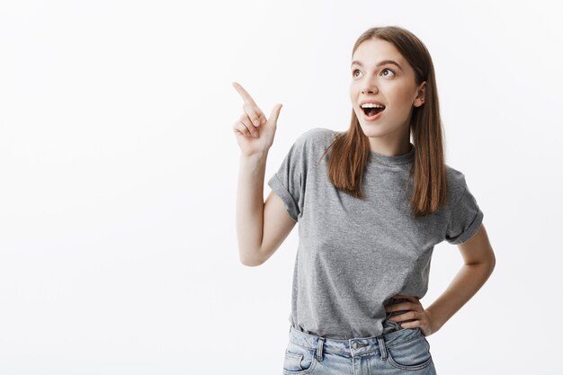 Retrato de menina atraente jovem estudante caucasiano com cabelo escuro em roupas casuais, olhando de lado, sorrindo com a boca aberta, apontando para a parede branca com o dedo. Copie o espaço.