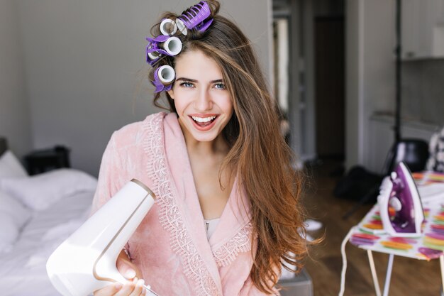 Retrato de menina bonita no roupão rosa com cachos na cabeça em casa. Ela segura o secador de cabelo, sorrindo.