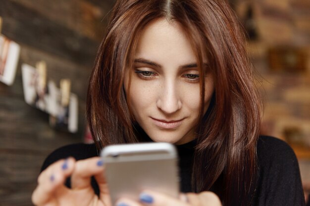 Retrato de menina bonita usando o touch screen celular lendo o artigo na revista on-line ou navegando na internet enquanto aguarda o cappuccino, descansando no restaurante sozinho. Foco seletivo no rosto