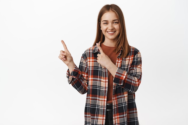 Retrato de menina caucasiana, sorrindo feliz, apontando os dedos no canto superior esquerdo, mostrando o texto promocional, copie o espaço para banner ou logotipo, em pé contra um fundo branco.