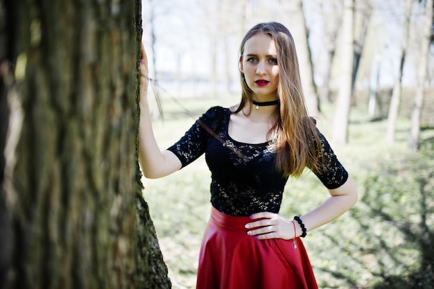 Retrato de menina com maquiagem brilhante com colar de gargantilha preta de lábios vermelhos no pescoço e saia de couro vermelha no parque primavera