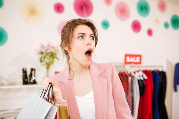 Retrato de menina de casaco rosa surpreendentemente olhando de lado com sacolas de compras na mão na loja de roupas senhora espantada em boutique com bolsos coloridos e cabideiro de venda no fundo