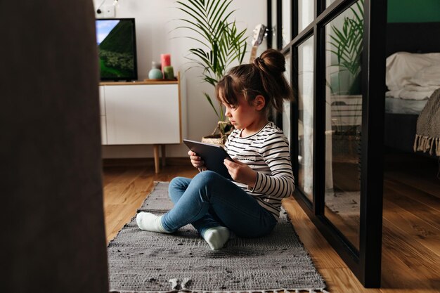 Retrato de menina de jeans sentado no tapete no quarto e jogando no tablet