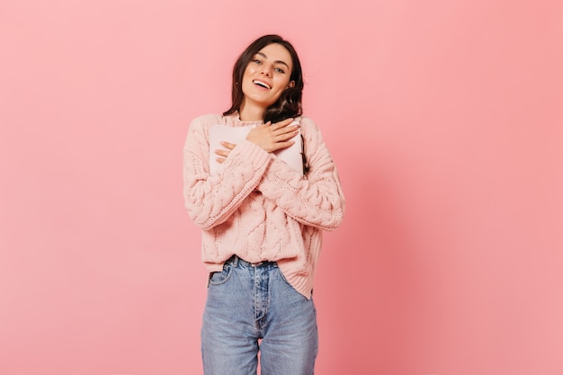 Retrato de menina de suéter rosa e jeans de mãe em fundo isolado. morena de olhos azuis com covinhas nas bochechas pressiona um diário importante contra o peito.