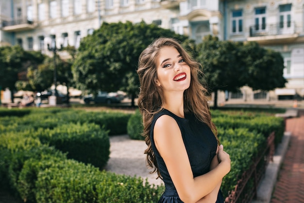 Retrato de menina elegante, com cabelos longos e lábios vínicos no pátio. ela usa vestido preto e sorri.