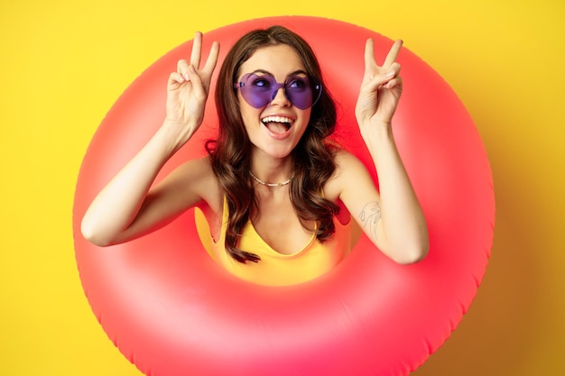 Retrato de menina elegante feliz em óculos de sol, dentro do anel de natação da praia, mostrando sinal de paz, rindo e sorrindo, de pé sobre fundo amarelo
