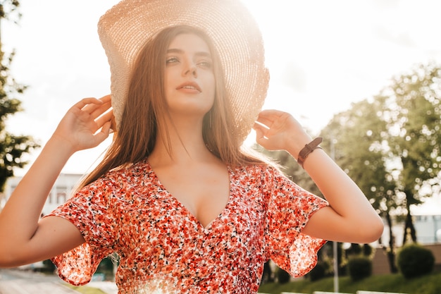 Retrato de menina jovem bonita hipster em vestido de verão na moda. mulher despreocupada sexy posando na rua fundo no chapéu ao pôr do sol. modelo positivo ao ar livre