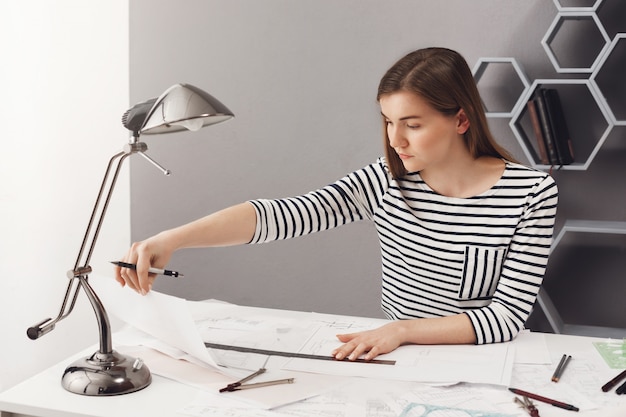 Retrato de menina jovem estudante morena com cabelos longos, camisa listrada, sentado à mesa em casa, fazendo o projeto de arquiteto para os exames, olhando desenhos com expressão concentrada no rosto.