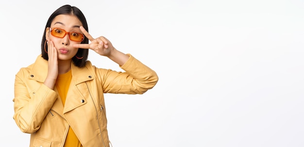 Retrato de menina moderna asiática elegante usando óculos escuros e jaqueta amarela mostrando gesto de paz vsign em pé sobre fundo branco feliz rosto sorridente