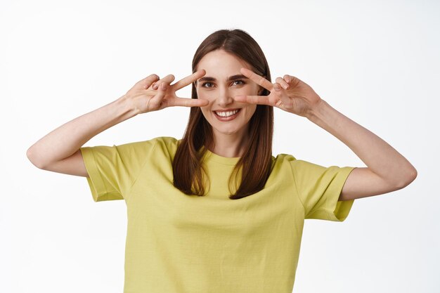 Retrato de menina morena positiva mostra gesto de sinal de paz perto dos olhos, enviando vibrações felizes, aproveitando as férias de verão, de pé na camiseta contra o fundo branco