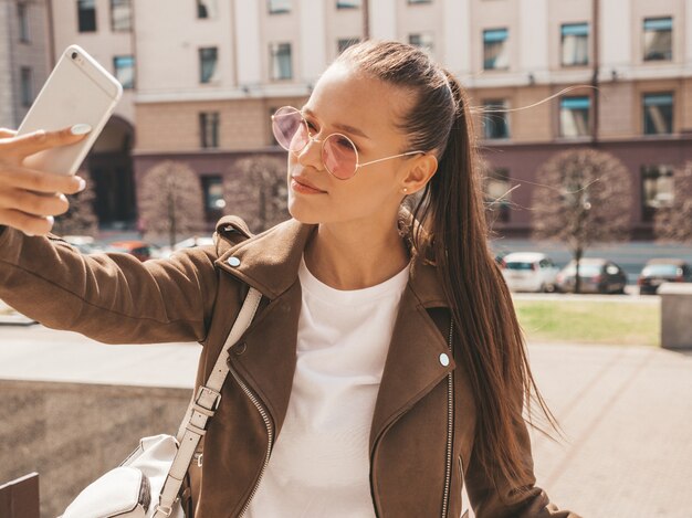 Retrato de menina morena sorridente linda jaqueta hipster de verão. Modelo tomando selfie no smartphone.