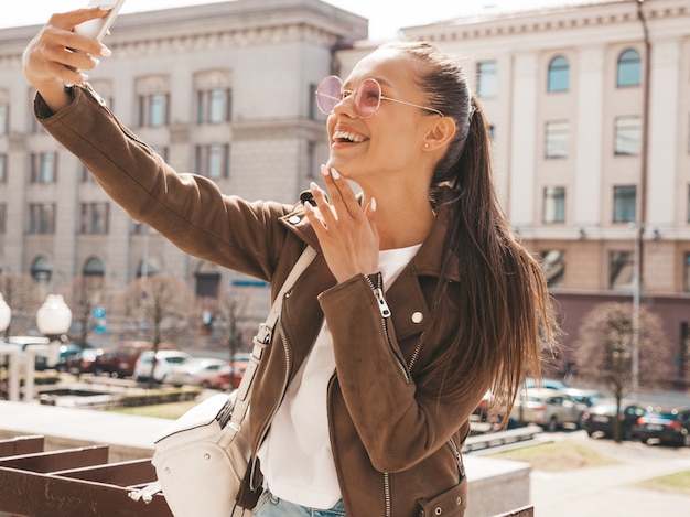 Retrato de menina morena sorridente linda jaqueta hipster de verão. Modelo tomando selfie no smartphone.