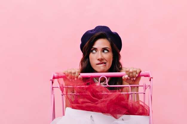 Retrato de menina na boina que adora fazer compras e segurando o carrinho. Mulher com cabelo preto encaracolado na boina posando em fundo rosa.