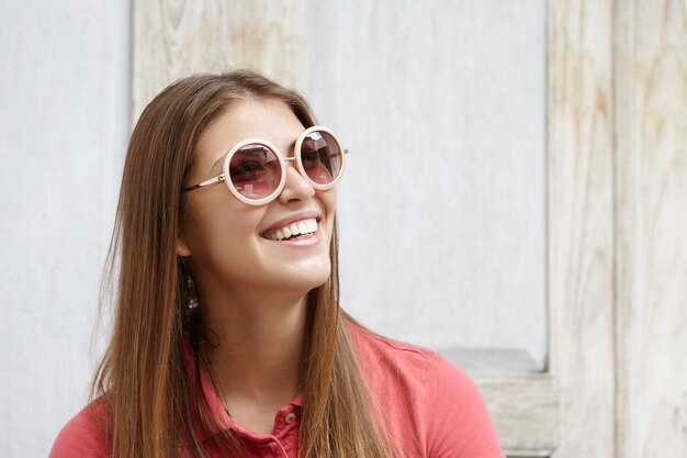 Retrato de menina na moda usando óculos de sol redondos com lentes espelhadas e camisa pólo, olhando para cima com um sorriso feliz, mostrando os dentes brancos, isolado contra a parede de madeira
