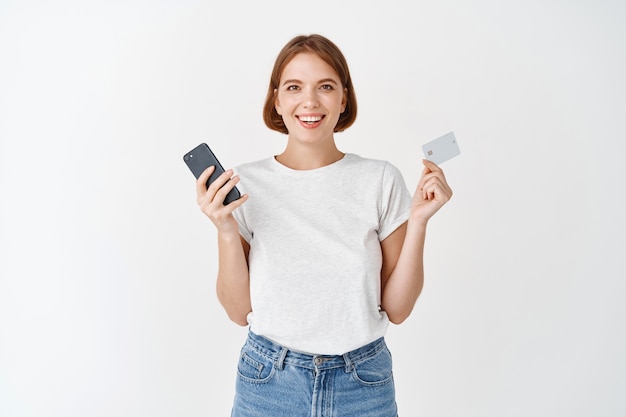 Retrato de menina natural sorridente mostrando telefone celular e cartão de crédito de plástico, pagamento online, em pé contra uma parede branca