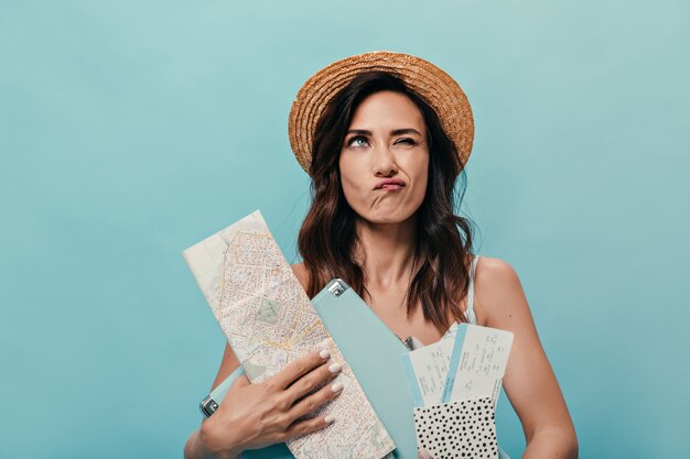 Retrato de menina posando pensativamente em fundo azul com o mapa da cidade e a mala. Mulher de chapéu de palha com cabelos escuros ondulados.