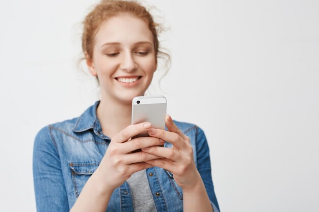 Retrato de menina ruiva encantadora positiva com penteado coque sorrindo sensualmente enquanto segura o smartphone e mensagens de texto