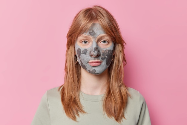 Retrato de menina ruiva séria de vinte anos olha diretamente para a câmera aplica máscara facial de argila para tratamento de pele vestida com camiseta casual isolada sobre fundo rosa Conceito de bem-estar