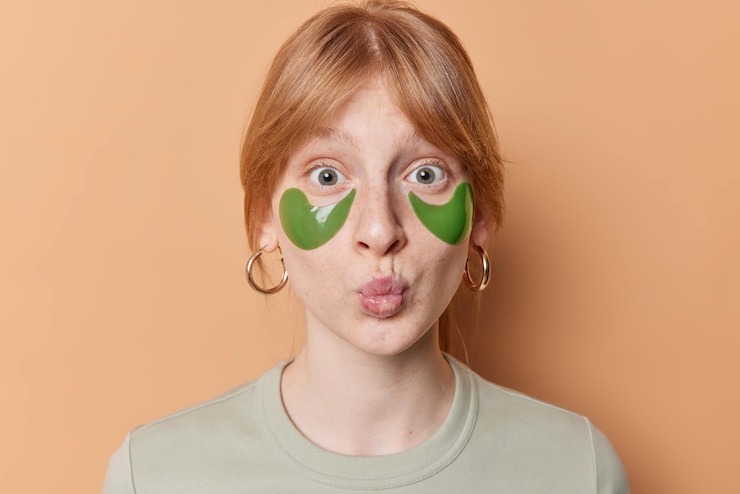 Foto grátis retrato de menina ruiva surpresa mantém os lábios dobrados aplica adesivos de colágeno verde para reduzir o inchaço sob os olhos usa poses de brincos contra fundo marrom cuidados com a pele das mulheres e conceito de beleza