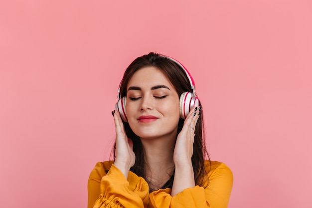 Retrato de menina satisfeita sem maquiagem em fones de ouvido na parede rosa. modelo sorrindo enquanto ouve uma melodia agradável.