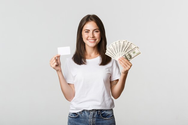 Retrato de menina sorridente animada segurando dinheiro e cartão de crédito, branco.