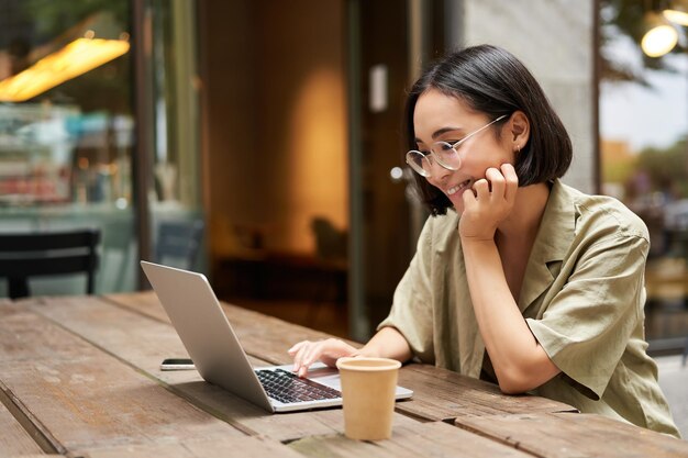 Retrato de menina sorridente em óculos sentado com laptop no café ao ar livre bebendo café e trabalhando