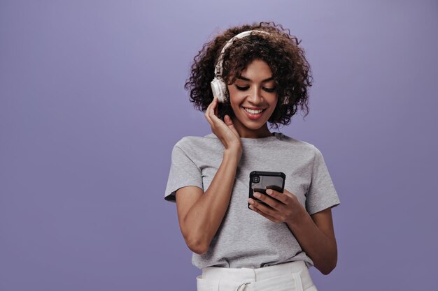 Retrato de menina sorridente, ouvindo música em fones de ouvido e segurando o telefone Mulher encaracolada feliz em camiseta cinza e shorts brancos conversando no celular no pano de fundo roxo