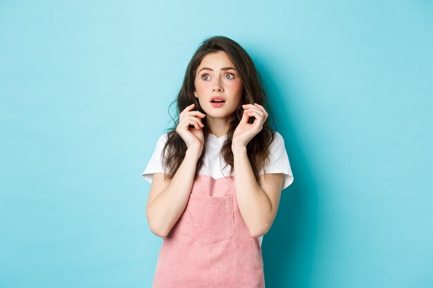 Retrato de menina tímida assustada com roupas de verão, olhando assustada para o banner de espaço de cópia, pulando de algo assustador, em pé contra um fundo azul.