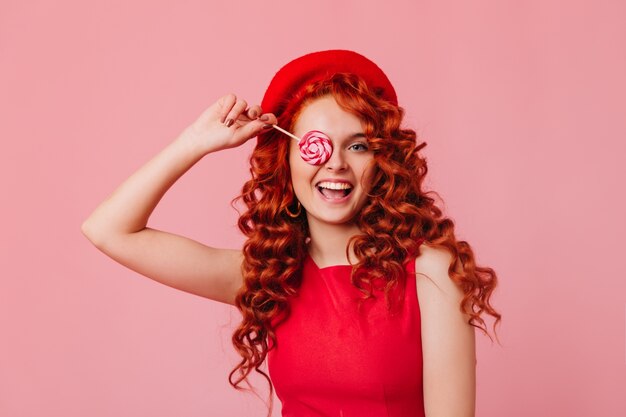 Retrato de menina travessa com cabelo ruivo ondulado no top brilhante e boina cobrindo os olhos com pirulito.