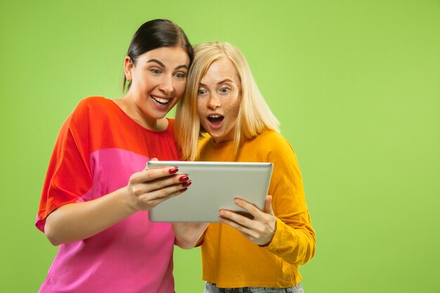 Retrato de meninas muito charmosas em roupas casuais, isoladas no fundo verde do estúdio. Namoradas ou lésbicas usando um tablet para se divertir ou pagamentos. Conceito de LGBT, emoções humanas, amor, relação.