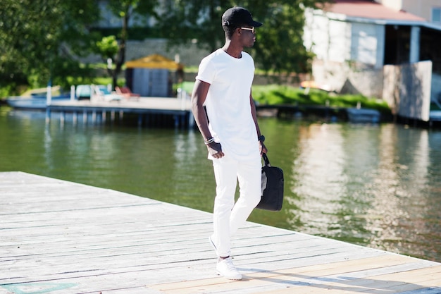 Retrato de menino afro-americano elegante usar óculos de roupas brancas e boné com bolsa na mão contra o cais do lago Moda de rua de jovens negros