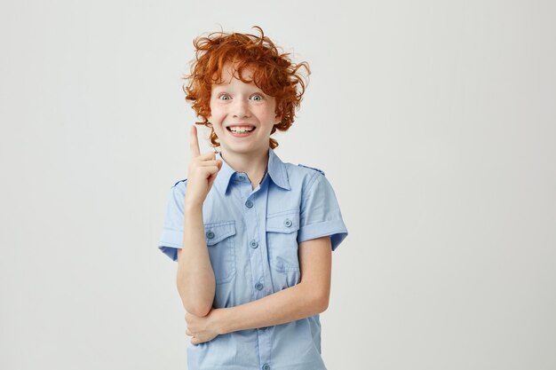 Retrato de menino alegre com cabelo ruivo e sardas sorrindo, apontando de cabeça com o dedo, tendo expressão louca. Copie o espaço.