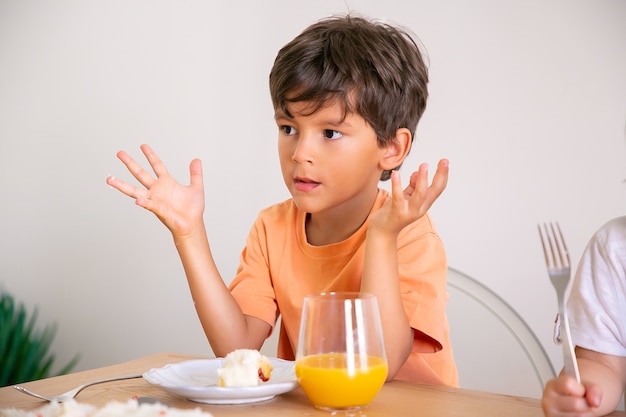 Retrato de menino bonitinho comendo bolo de aniversário e bebendo suco de laranja. Adorável criança sentada à mesa na sala de jantar, levantando as mãos e olhando para longe. Conceito de infância, celebração e férias