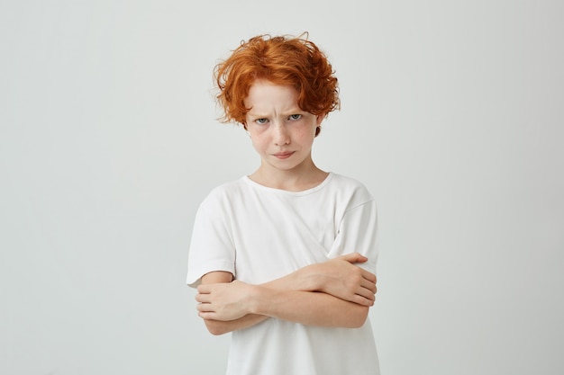 Retrato de menino de cabelo vermelho infeliz com sardas olhando com expressão chateada, cruzando as mãos sendo insatisfeito que sua mãe o repreendeu.