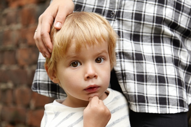 Retrato de menino de olhos castanhos e sua mãe. criança de cinco anos com cabelo loiro em pé quieto. sua mãe amorosa parada perto, acariciando sua cabeça. expressão facial curiosa.