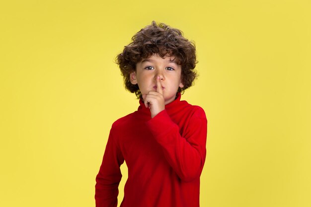 Retrato de menino muito jovem encaracolado no desgaste vermelho sobre fundo amarelo do estúdio. Infância, expressão, educação, conceito divertido. Pré-escolar com expressão facial brilhante e emoções sinceras.
