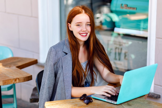 Retrato de moda ao ar livre da cidade de jovem empresária trabalhando no café no terraço em dia de sol, roupa elegante casual, detalhes de hortelã, usando seu laptop, pausa para café, conceito de negócio.