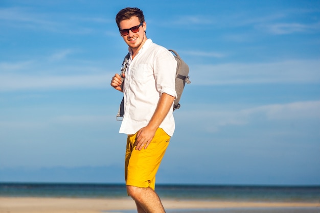 Retrato de moda ao ar livre de homem bonito posando na incrível praia tropical, em um belo dia de sol, bela vista do céu azul e do oceano, vestindo óculos escuros e camisa branca clássica de tipo amarelo casual.