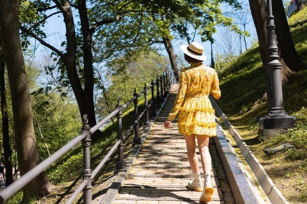 Retrato de moda ao ar livre de mulher com vestido amarelo de verão e chapéu andando no caminho na vista traseira do parque