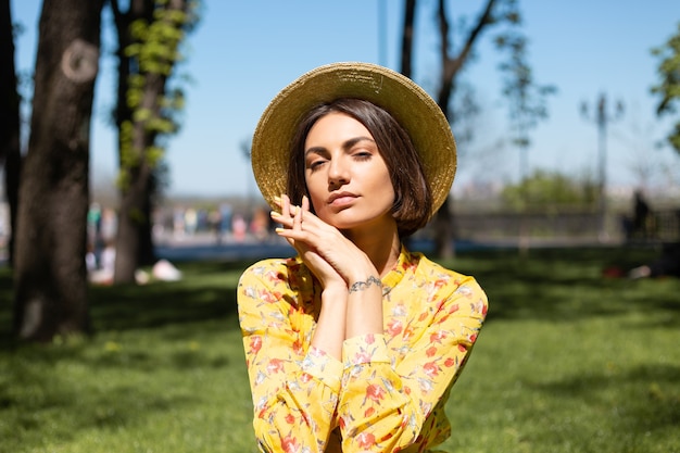 Retrato de moda ao ar livre de mulher com vestido amarelo de verão e chapéu sentada na grama do parque