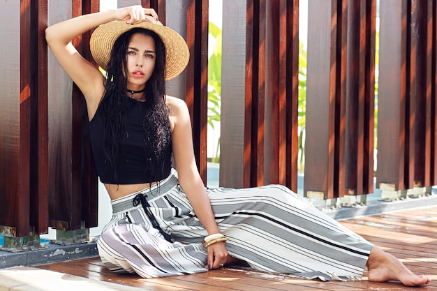 Retrato de moda ao ar livre de mulher sensual com chapéu de palha, calças listradas elegantes posando em villa de luxo perto da piscina