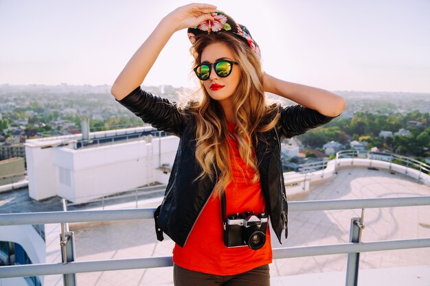 Retrato de moda ao ar livre de uma garota fotógrafa elegante segurando uma câmera retro vintage, usando um chapéu brilhante, óculos de sol da moda e jaqueta de couro, vista incrível da cidade do telhado