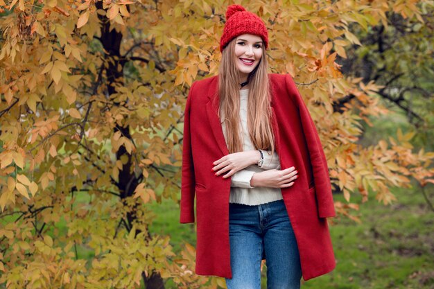 Retrato de moda outono de mulher loira feliz casaco vermelho elegante e chapéu de malha andando no parque.