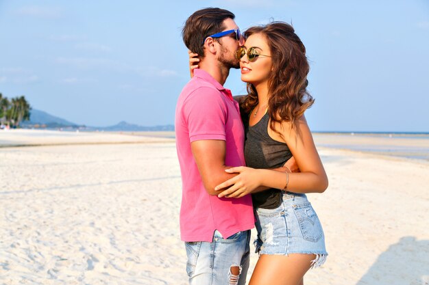 Retrato de moda verão de jovem casal de descolados muito elegantes, abraços de amor e posando na incrível praia da ilha, se divertindo sozinho, usando óculos escuros e roupas casuais brilhantes.