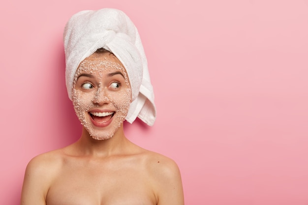 Retrato de modelo feminina feliz aplica esfoliante de sal marinho no rosto, tem expressão positiva, olha para o lado, está nu, usa toalha após o banho