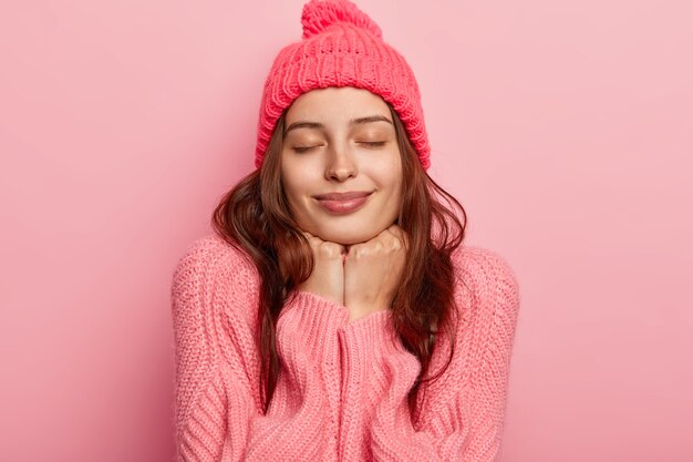 Retrato de modelo feminino jovem e relaxado mantém as duas mãos sob o queixo, tem os olhos fechados, usa macacão e chapéu rosado, sente-se satisfeito, posa contra um fundo rosa.