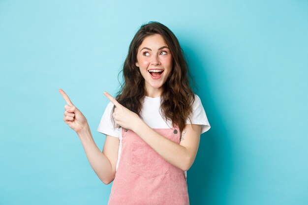 Retrato de modelo feminino sorridente e feliz com blush de glamour, apontando o dedo para a esquerda e olhando para a oferta promocional, mostrando um espaço de cópia de anúncio, fundo azul