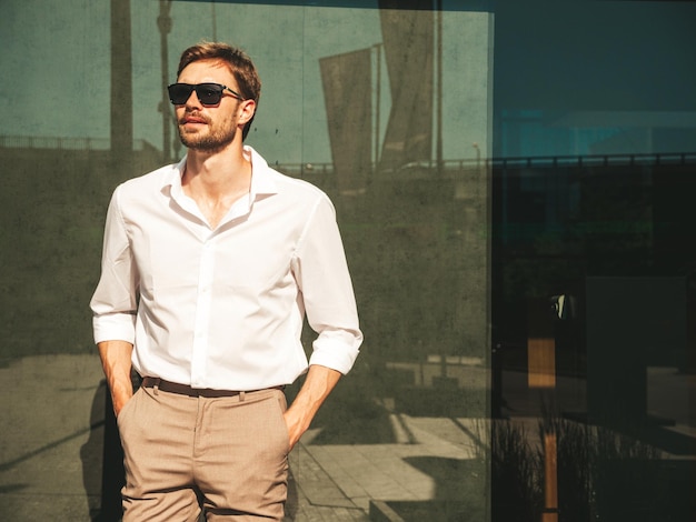 Retrato de modelo lambersexual bonito confiante elegante hipster homem moderno sexy vestido com camisa branca e calças moda masculina posando no fundo da rua na cidade da europa ao pôr do sol em óculos de sol