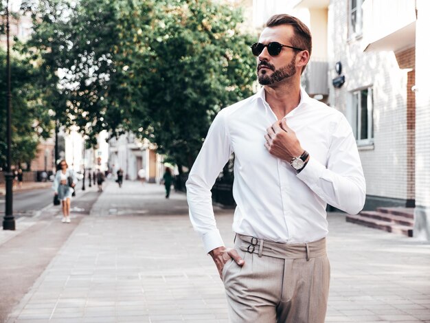 Retrato de modelo lambersexual bonito confiante elegante hipster homem moderno sexy vestido com camisa branca e calças Moda masculina posando no fundo da rua na cidade da Europa ao pôr do sol em óculos de sol