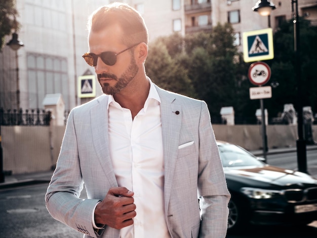 Retrato de modelo lambersexual bonito confiante elegante hipster homem moderno sexy vestido com terno branco elegante moda masculina posando no fundo da rua na cidade da europa ao pôr do sol em óculos de sol